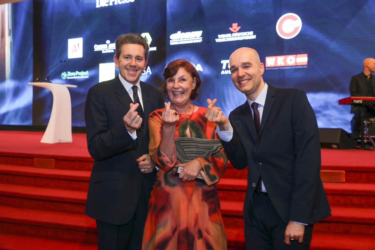 Harald Mahrer, Karin Simonitsch und Sreco Dolanc stehen vor einer Bühne und zeigen die Gebärde für Liebe