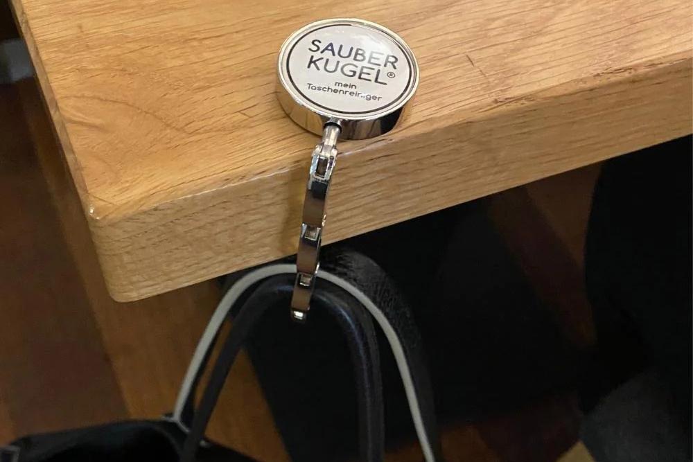 Taschenhalter von Sauberkugel, der an eine Tischkante geklemmt ist. Daran hängt eine schwarze Ledertasche, von der nur der Griff zu sehen ist.