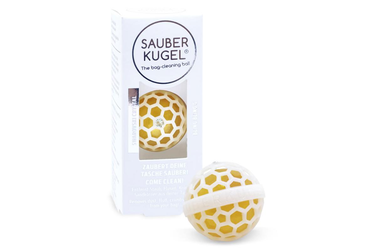 Sauberkugel in der weißen Schachtel. Davor liegt eine lose goldene Kugel mit weißem Gehäuse und Swarovski-Kristall.