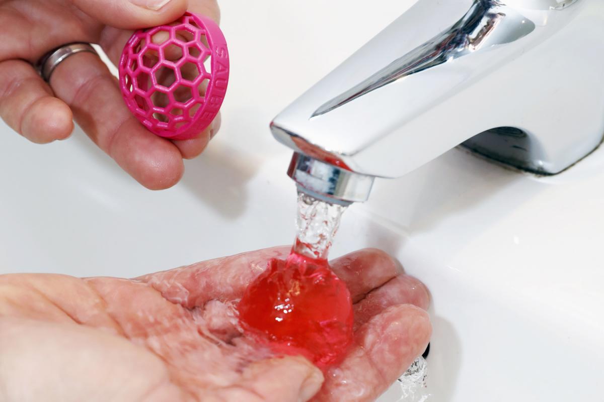 Pinke Sauberkugel, die in einem Waschbecken unter fließendes Wasser gehalten wird. In der anderen Hand befindet sich die Hülle der Sauberkugel.