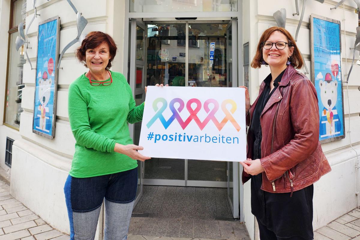 Mag. Karin Simonitsch und Andrea Brunner, Geschäftsführerin der Aids Hilfe Wien, stehen vor der Marien Apotheke in 1060 Wien. Sie halten gemeinsam ein Schild mit der Aufschrift