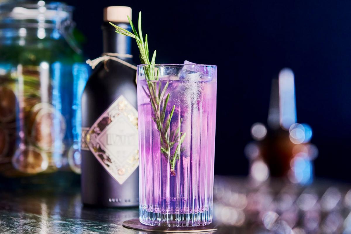Eine schwarze Flasche des alkoholfreien Gins The Illusionist Masquerade. Daneben ein Glas mit rosa Gin Tonic und Orangenschale, das von einem Barkeeper auf den Tisch gestellt wird.