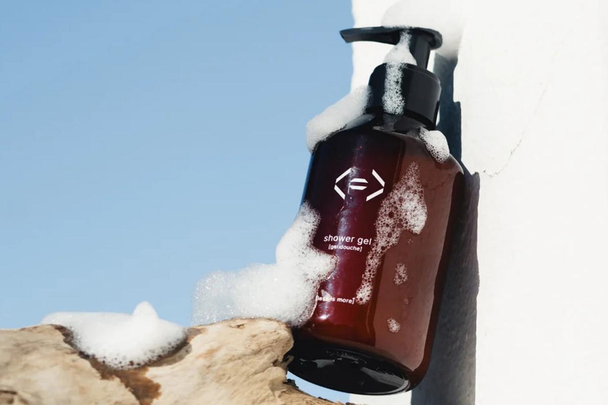 Duschgel in brauner Glasflasche von Less is More. Es lehnt an einer Mauer und steht auf einem Stück Treibholz