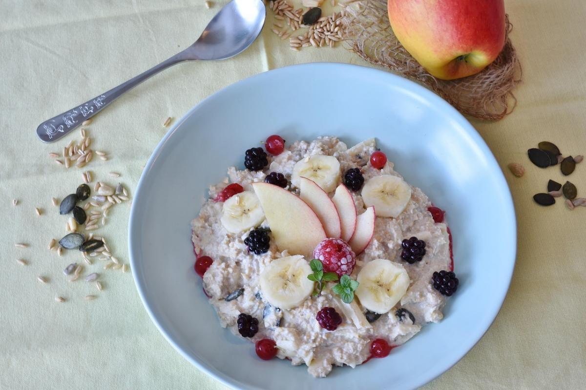 Porridge aus Haferflocken mit Bananen und Heidelbeeren. Daneben liegen ein Löffel und ein Apfel.