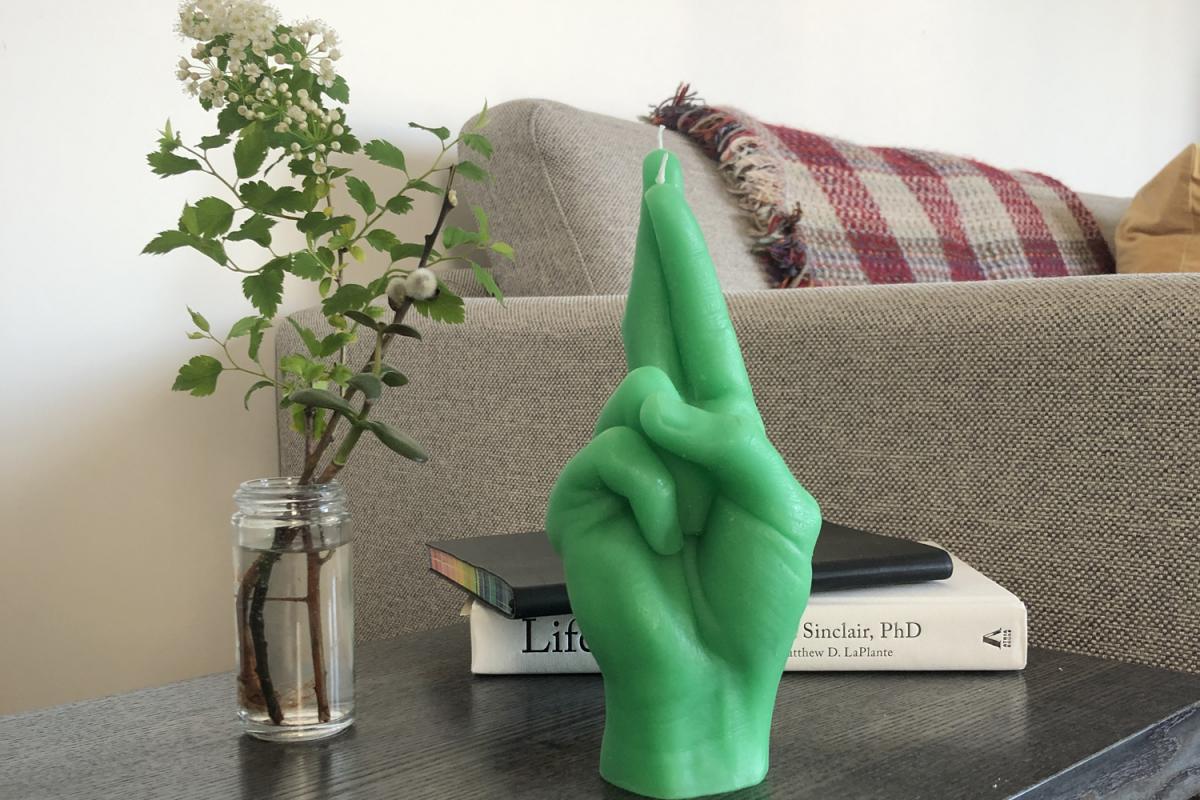 Grüne Kerze in Handform, deren Finger verkreuzt sind. Die Kerze steht auf einem Tisch vor einem Buch und neben einer Vase mit Blumen.